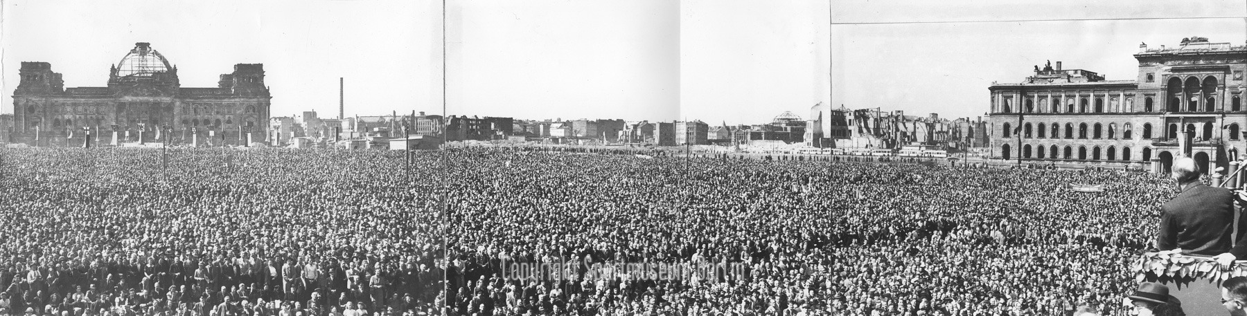 Schwarz-Weiß-Fotos montiert: Versammlung von tausenden Menschen. Im Vordergrund rechts Redner vor Mikrofonen. Im Hintergrund zerbombte Häuser, u. a. der Reichstag.