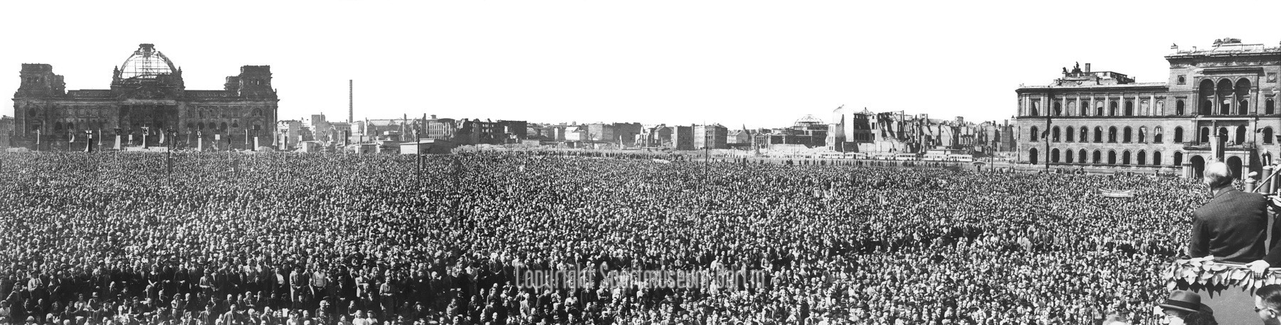 Schwarz-Weiß-Foto: Versammlung von tausenden Menschen. Im Vordergrund rechts Redner vor Mikrofonen. Im Hintergrund zerbombte Häuser, u. a. der Reichstag.